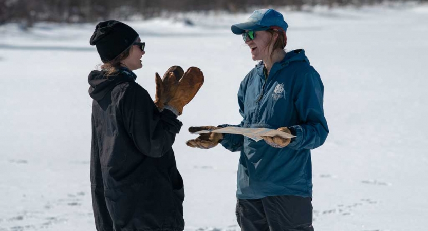 Two people wearing winter gear talk to each other in a snowy landscape 